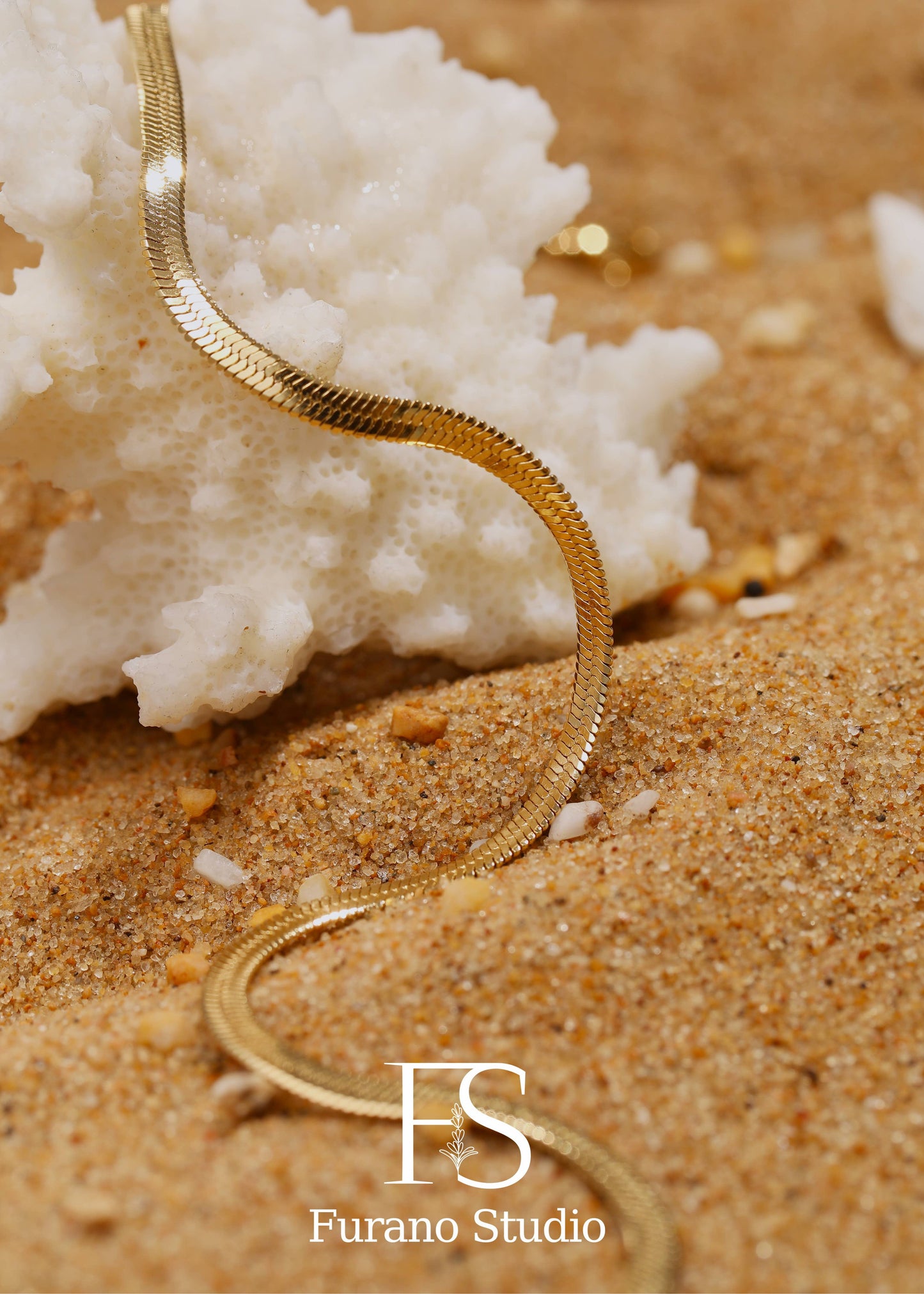 Gold necklace on sand with white coral, Furano Studio logo.