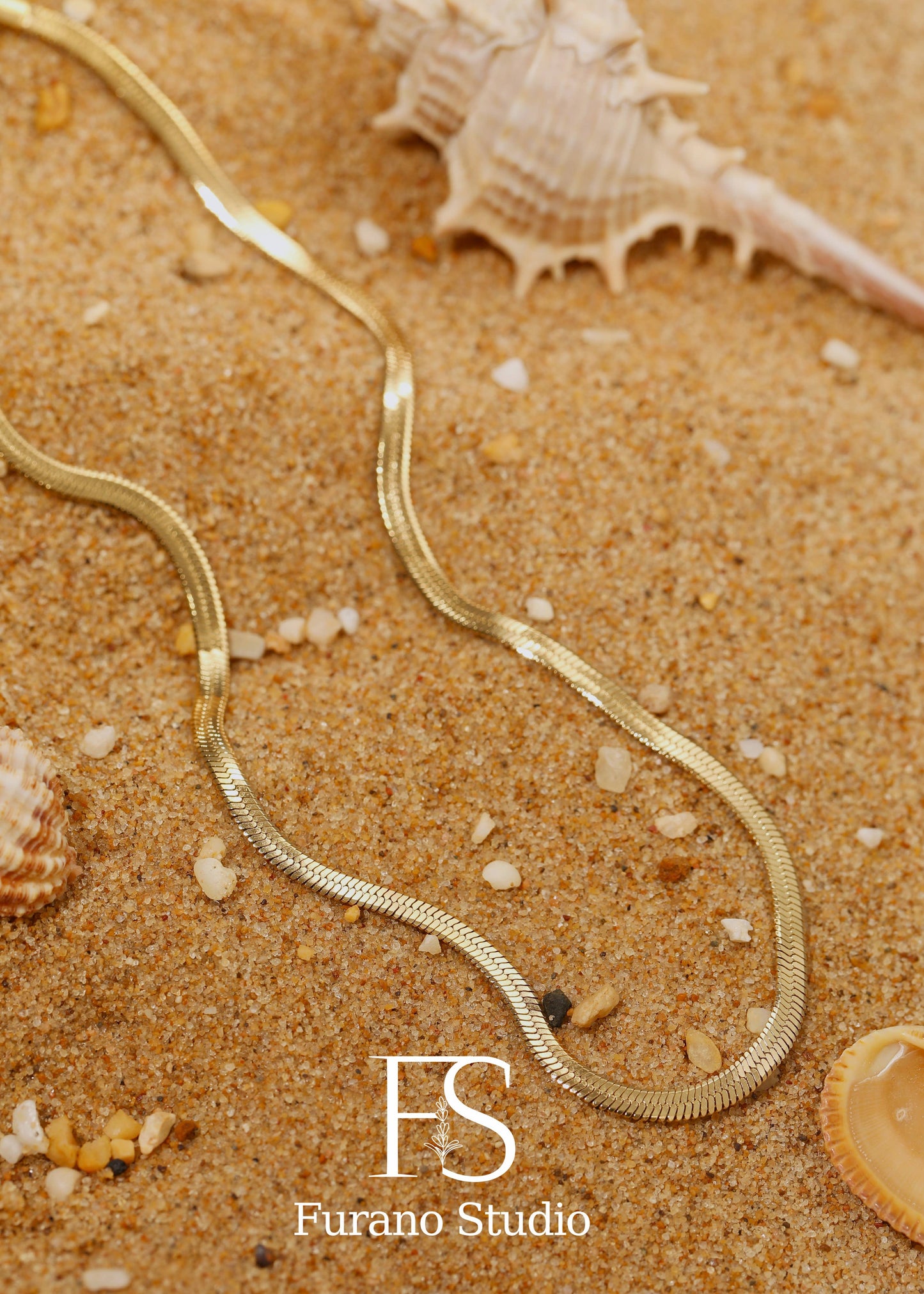 Gold necklace on sand with seashells, featuring Furano Studio logo.