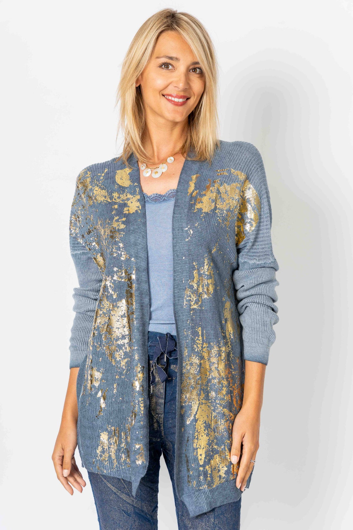 A woman modeling a blue open-front cardigan with a distressed gold foil pattern, worn over a light blue top and blue jeans against a light gray background.