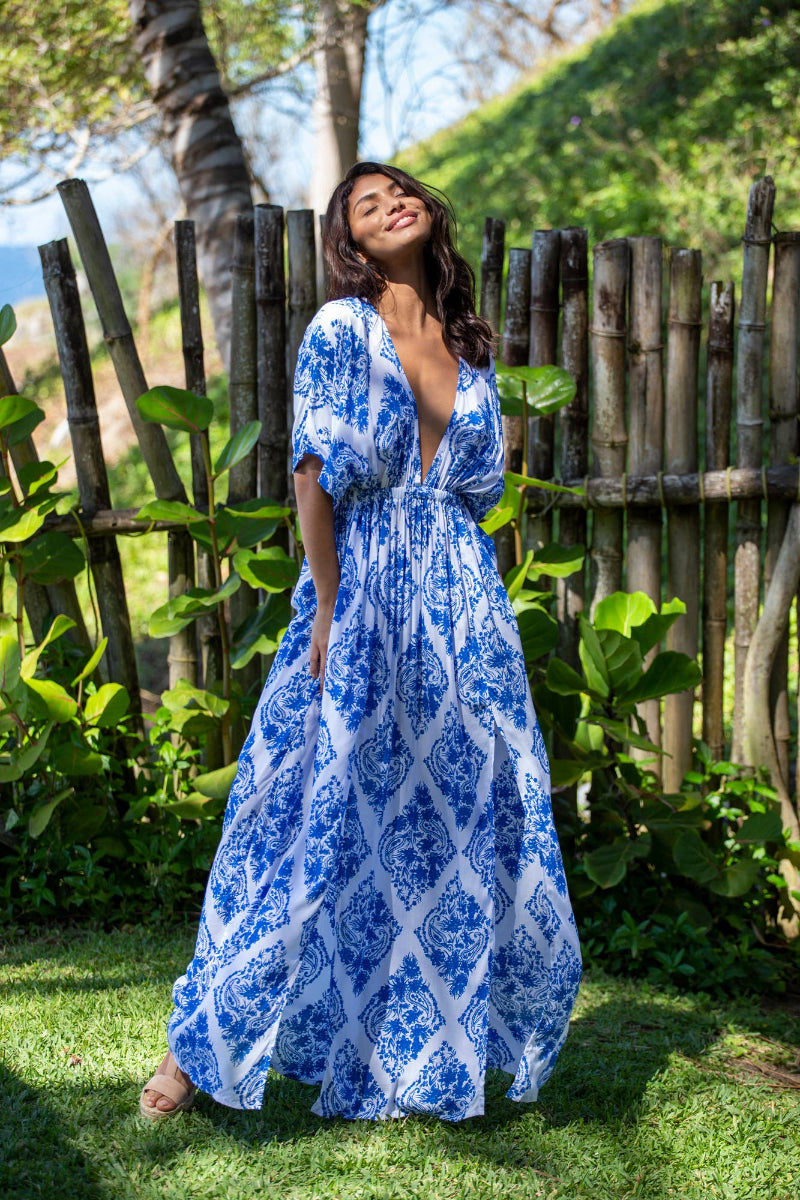 Woman in a blue and white patterned dress standing outdoors with greenery and a wooden fence in the background.