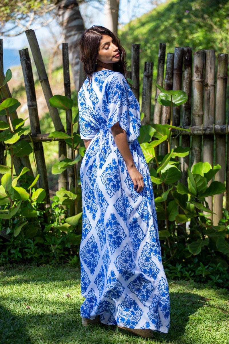 Woman in a blue and white patterned dress standing in a garden with greenery and wooden fence.