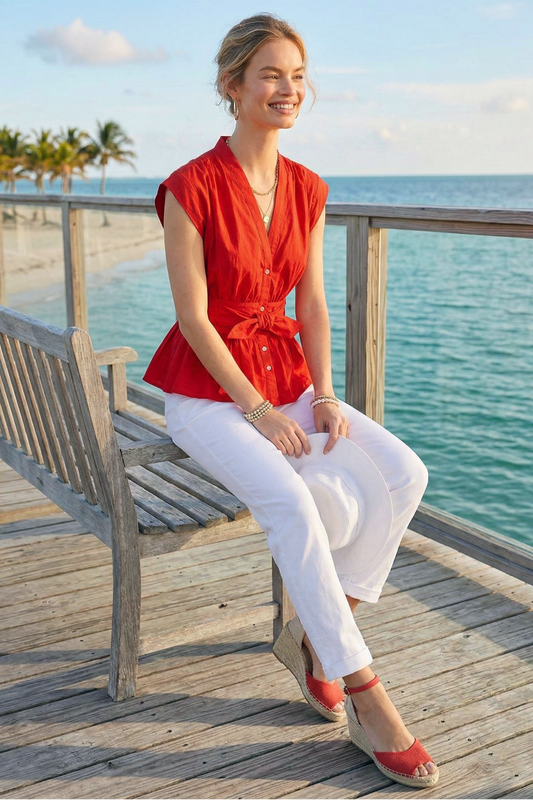 Woman in a red top and white pants sitting on a wooden bench by the ocean.