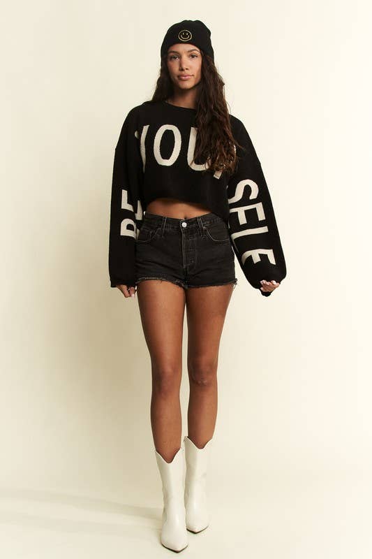 Person wearing a black sweater with text, denim shorts, and white boots on a plain background