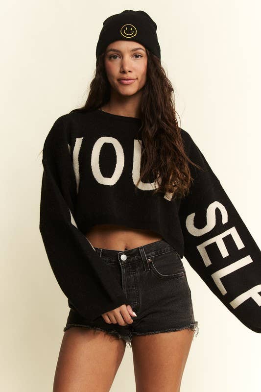 Person wearing a black sweater with 'YOU SELF' text, black beanie, and black shorts on a beige background