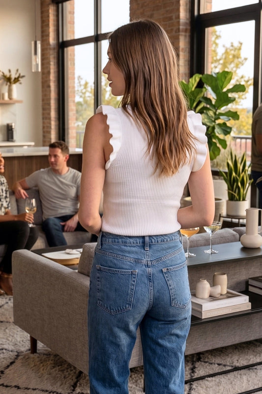 Woman in a white top and blue jeans standing in a modern living room with a man sitting on a couch.