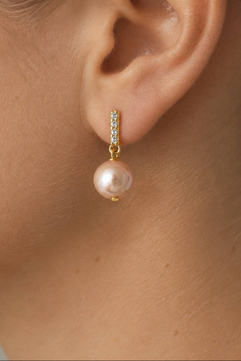 A close-up of a model's ear wearing a gold huggie hoop earring with a vertical row of small clear stones and a dangling pink pearl.
