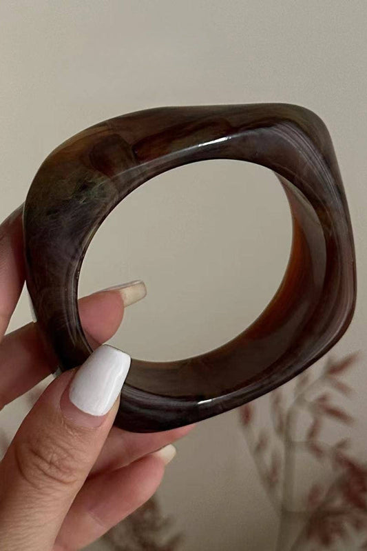 Brown bangle held by a hand with white nail polish against a neutral background