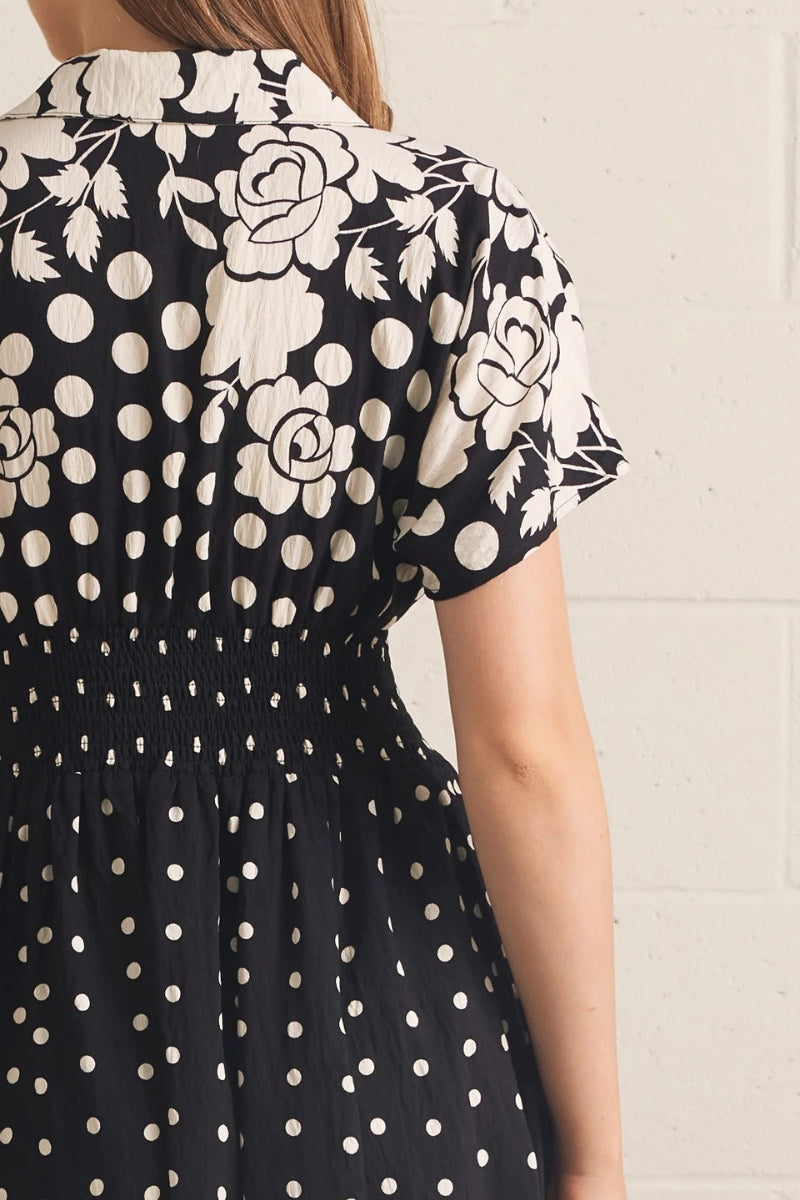 A close-up back view of a woman wearing a black dress with a white floral rose print on the upper back and shoulders, and a white polka dot pattern on the skirt, against a light brick wall.