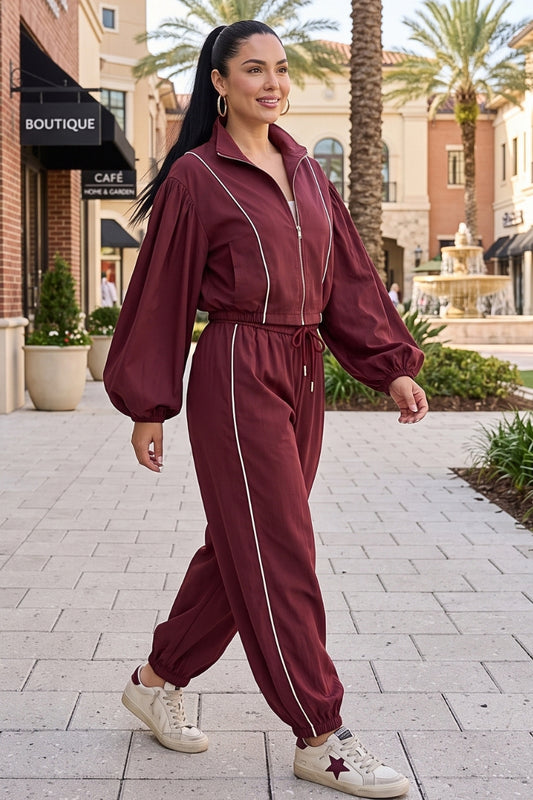 A woman modeling a maroon zip-up tracksuit with white piping and drawstrings, walking on a paved outdoor shopping plaza with palm trees and boutique storefronts in the background.