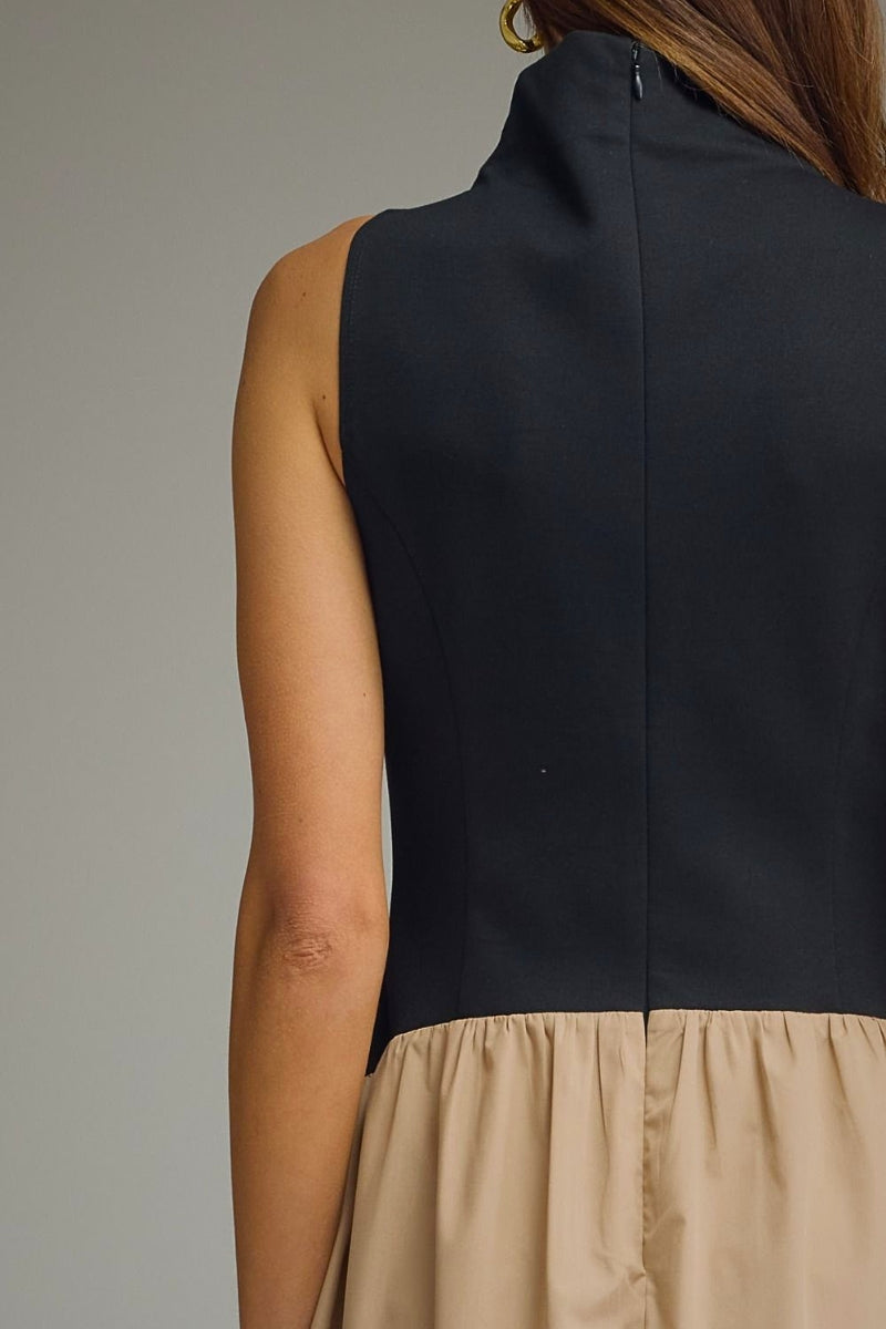 Close-up back view of a sleeveless dress with a high mock neck, black upper section, and beige gathered skirt, shown on a model against a plain gray background.