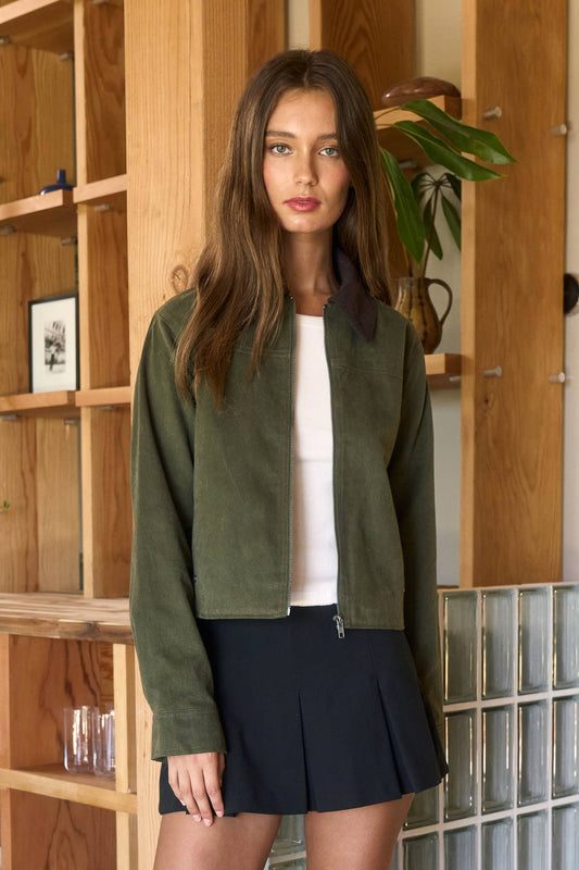 Woman wearing a green jacket and black skirt standing in a room with wooden shelves and a plant.