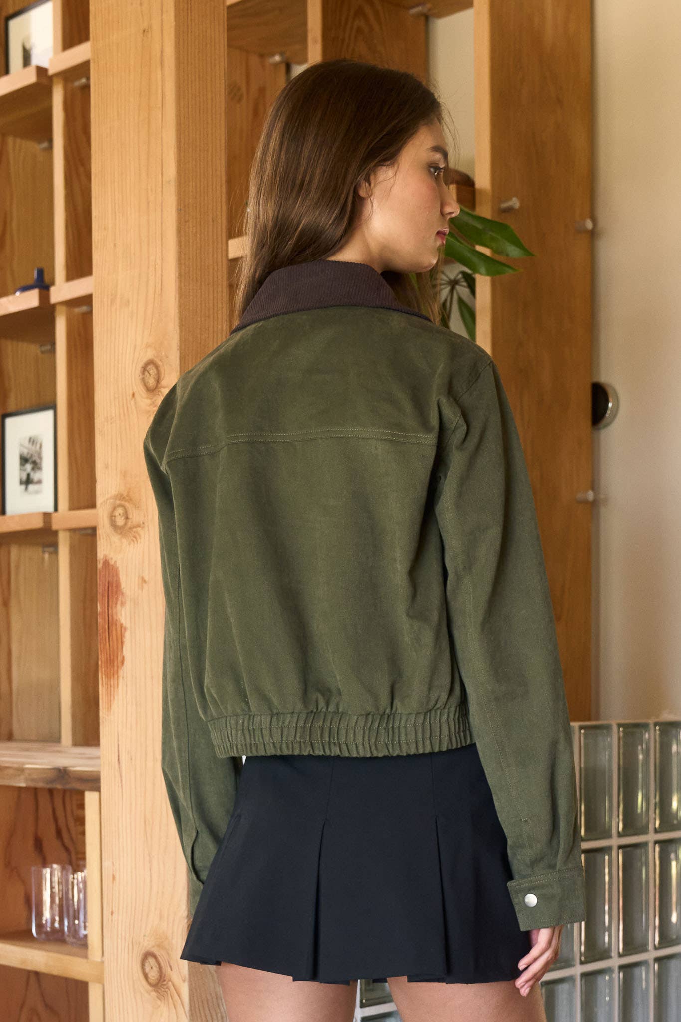 Woman wearing a green jacket and black skirt standing in a room with wooden shelves and a tiled wall.