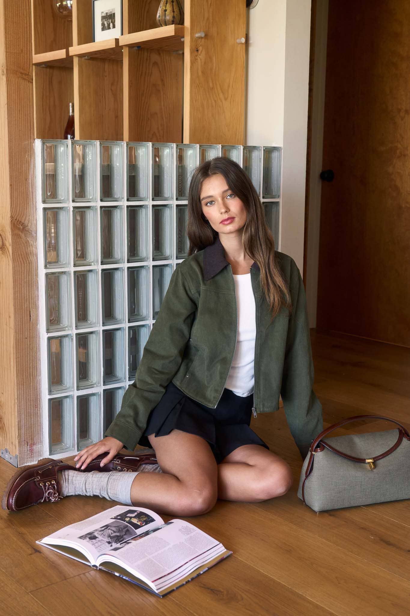Woman sitting on the floor with a book and a handbag in a room with wooden shelves and glass block wall.