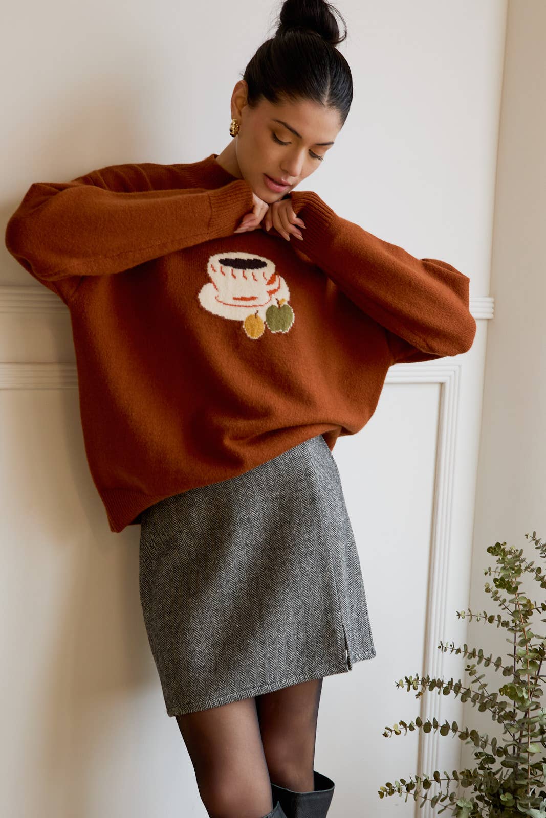 Woman wearing a brown sweater with a coffee cup design, standing against a white wall.