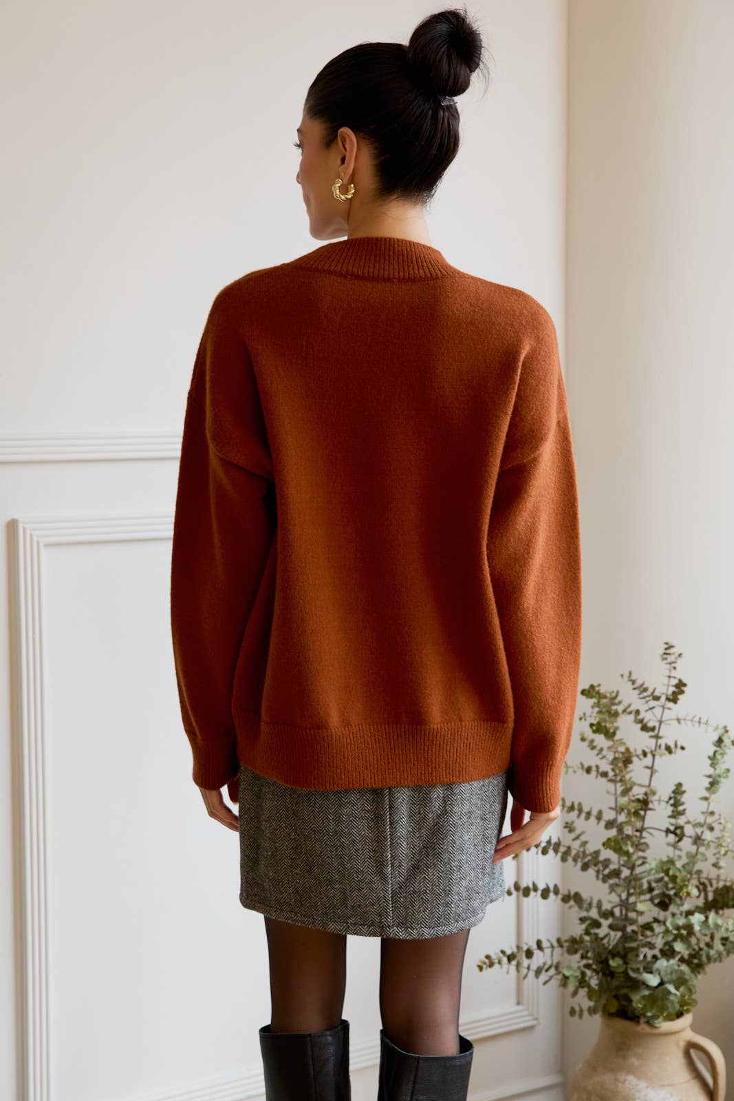 Person wearing a brown sweater and gray skirt in a room with a white wall and plant.
