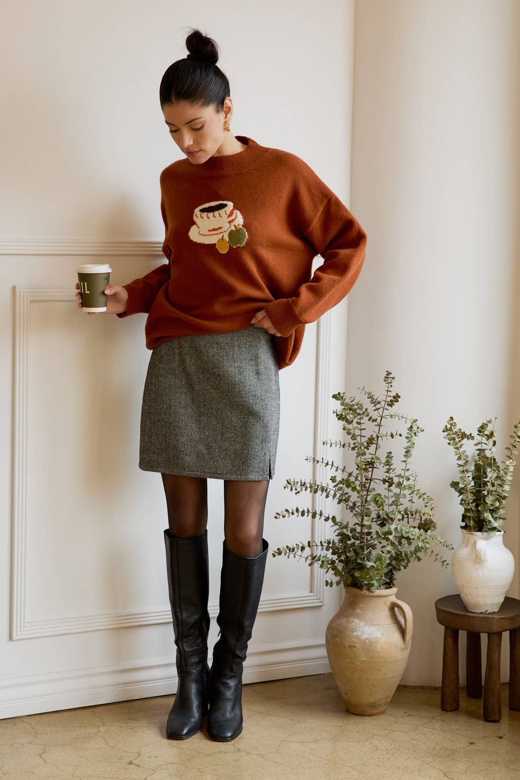 Woman wearing a brown sweater with a coffee design, gray skirt, and black knee-high boots, holding a mug.