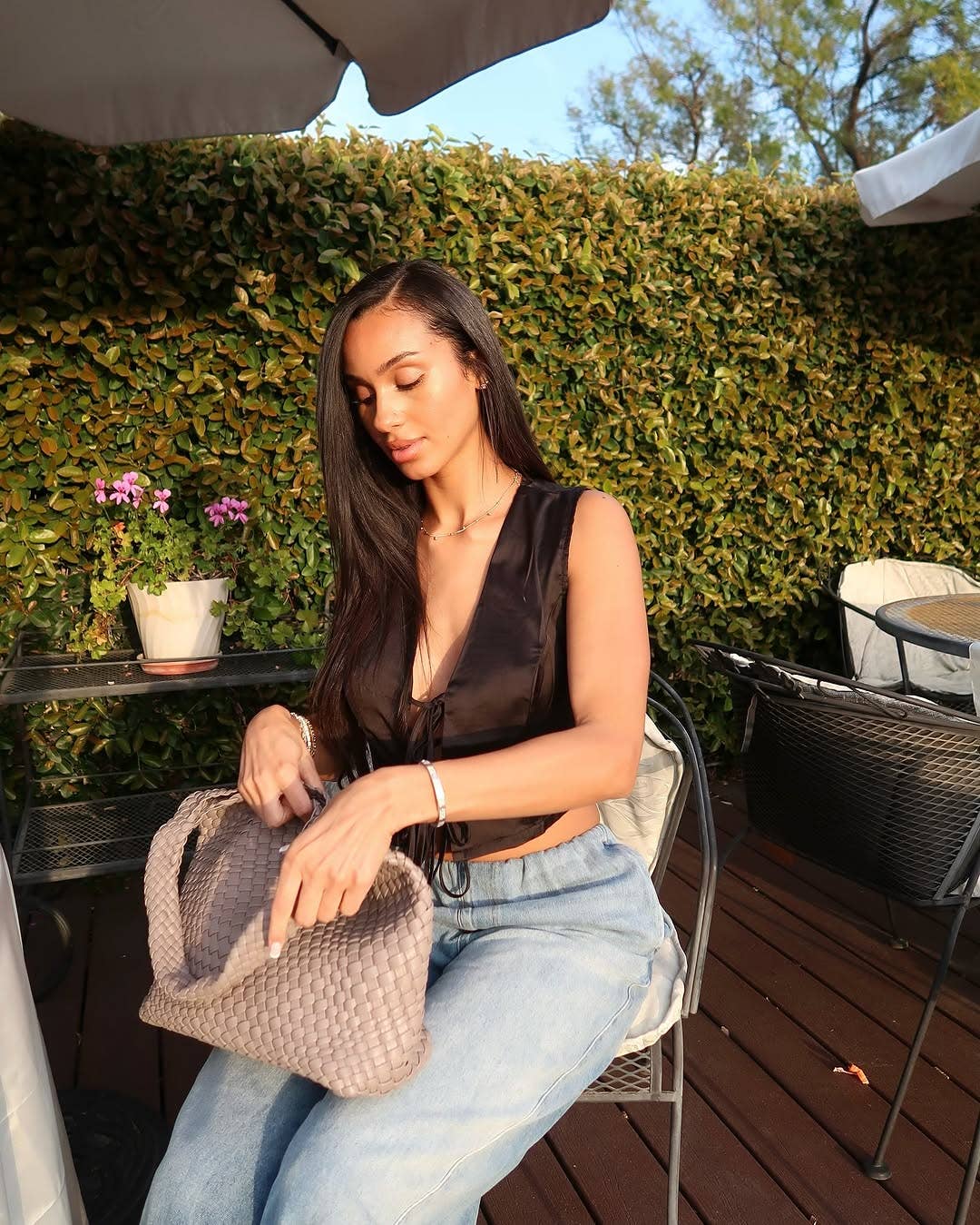 Woman sitting outdoors holding a woven handbag with greenery and tables in the background