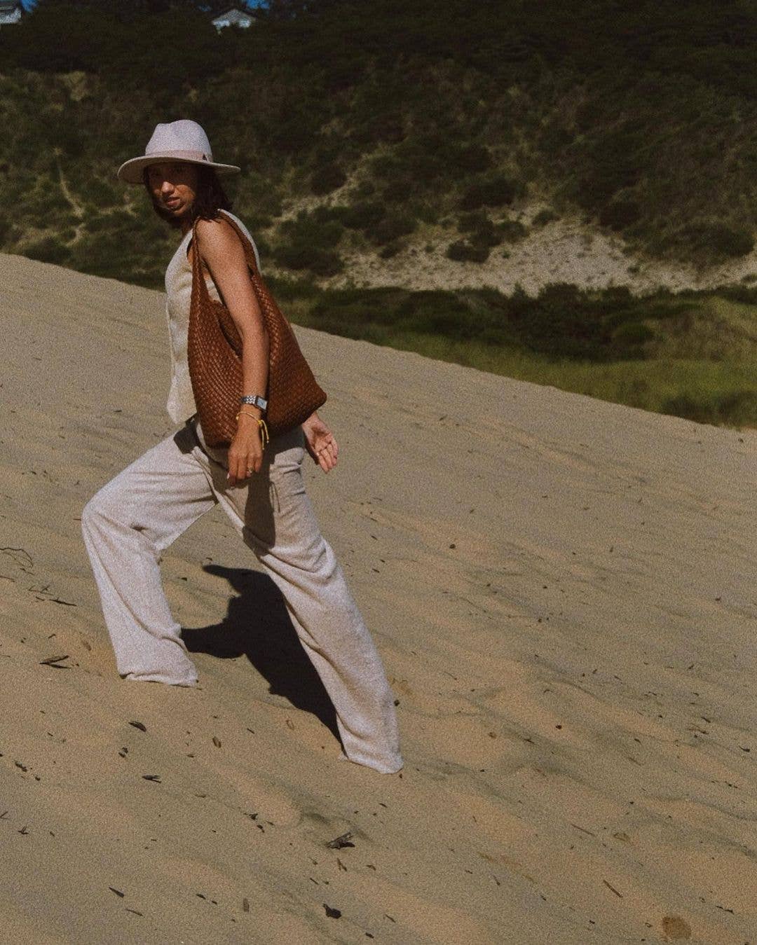 Woman walking on a sandy dune wearing a hat, beige outfit, and saddle bag.