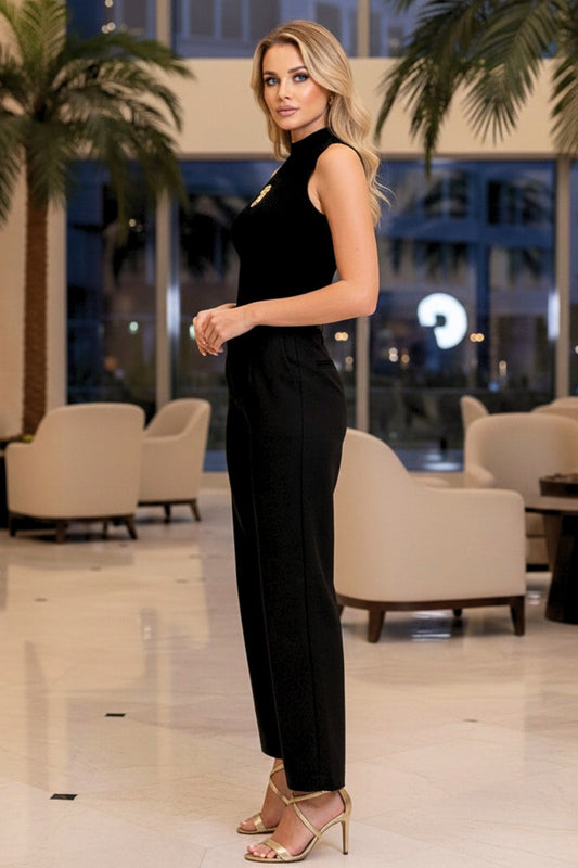 Woman in a black jumpsuit standing in an indoor setting with palm trees and chairs.