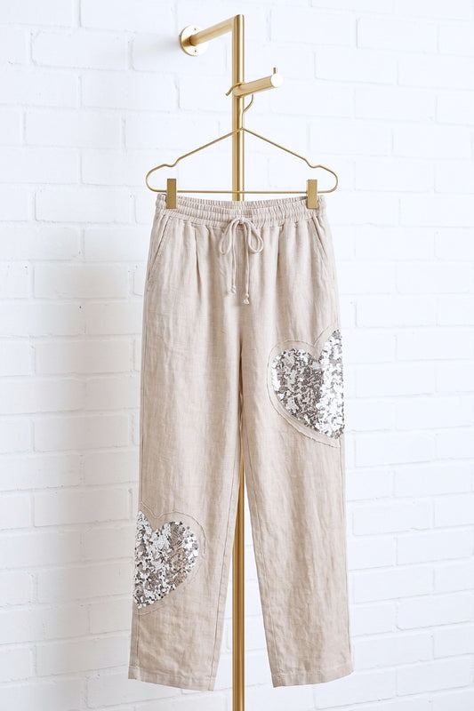 Beige pants with floral pocket details hanging on a gold rack against a white brick wall.