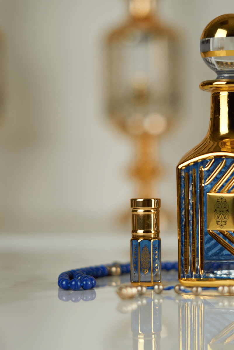 An ornate gold and blue perfume decanter with a matching small atomizer bottle and blue beaded bracelet, displayed on a reflective white surface with a warm, blurred background.