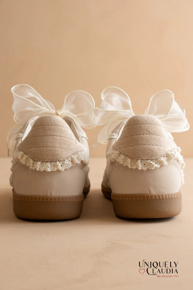 Pair of white shoes with lace trim and ribbons on a beige background, featuring the brand 'Uniquely Claudia'.