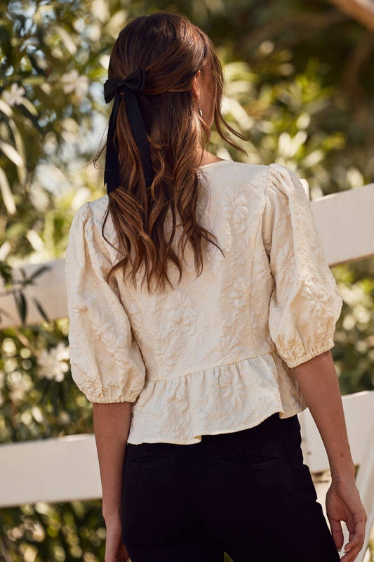 Woman wearing a beige lace blouse with black pants, standing outdoors with greenery in the background.