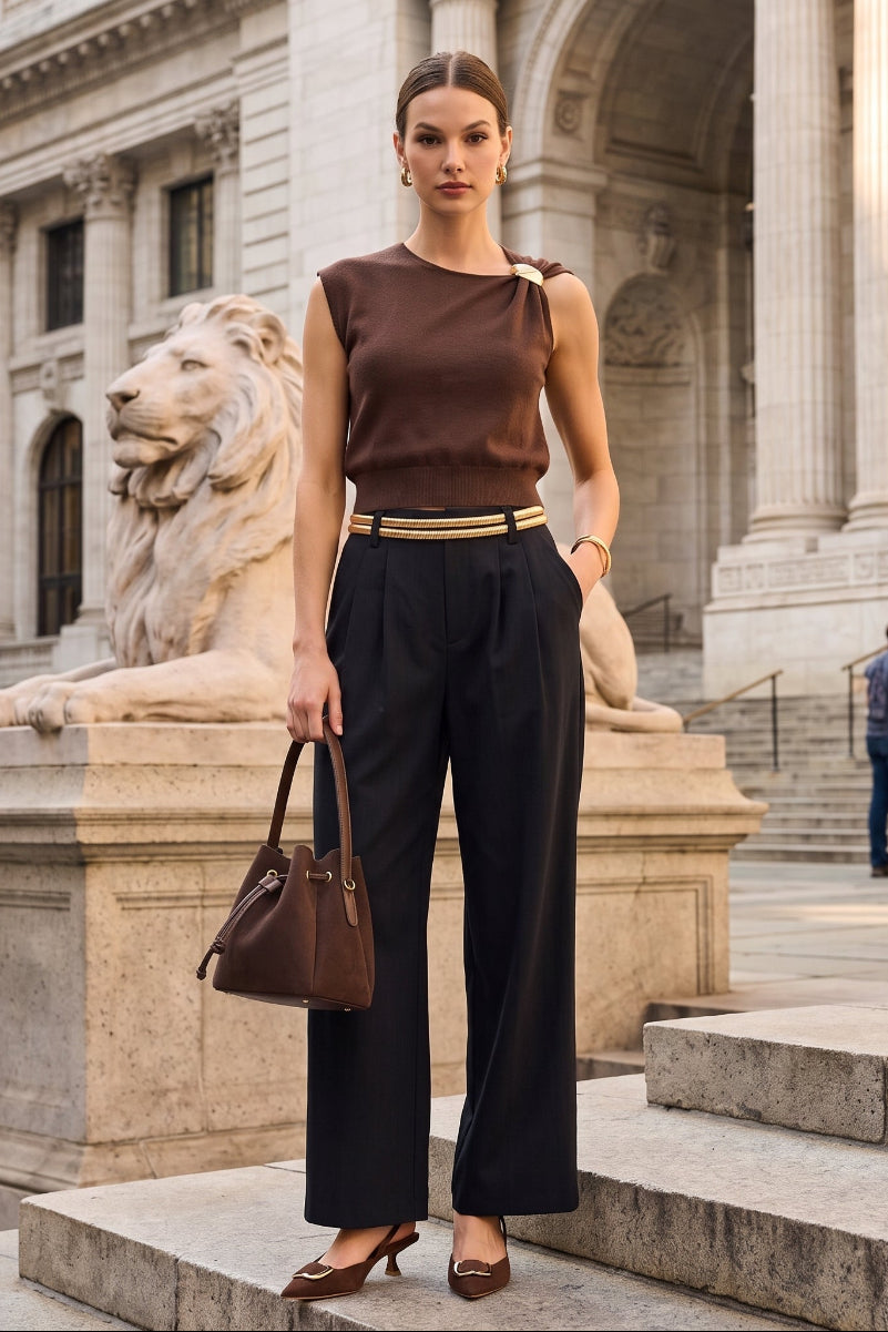 A woman stands on stone steps in front of a grand classical building, wearing a brown sleeveless top, black wide-leg trousers, and brown heels, while holding a brown handbag.