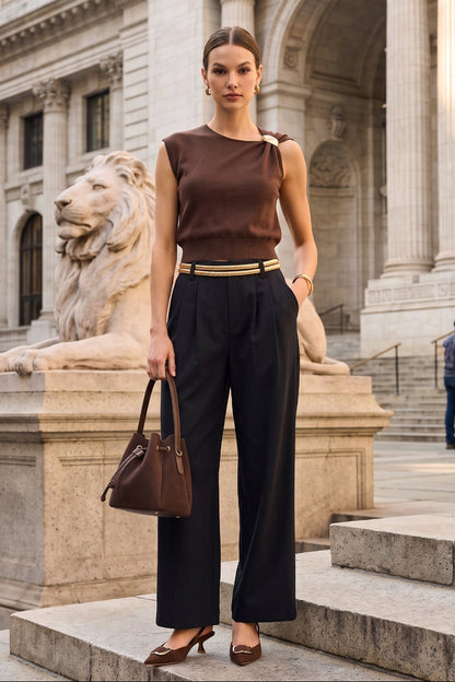 A woman stands on stone steps in front of a grand classical building, wearing a brown sleeveless top, black wide-leg trousers, and brown heels, while holding a brown handbag.