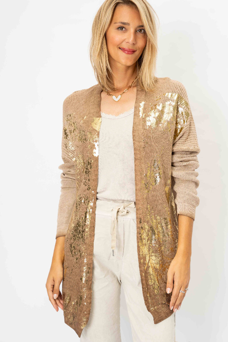 A woman modeling a long open-front cardigan covered in shimmering gold sequins over a taupe knit base, worn over a white top and white drawstring pants against a plain white studio background.