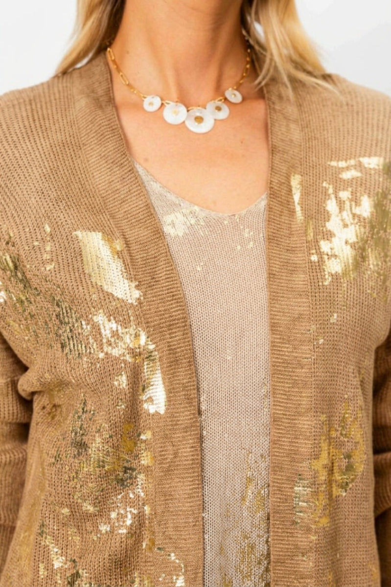 A close-up of a woman wearing a textured tan knit cardigan with gold sequin accents over a matching beige top, accessorized with a white shell necklace.