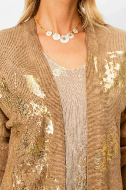 A close-up of a woman wearing a textured tan knit cardigan with gold sequin accents over a matching beige top, accessorized with a white shell necklace.