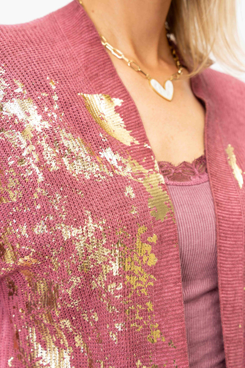 A close-up of a person wearing a textured pink knit cardigan with metallic gold leaf patterns, layered over a mauve ribbed top and a gold heart pendant necklace.