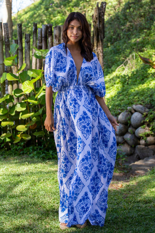 Woman in a blue patterned dress standing outdoors with greenery and wooden elements in the background