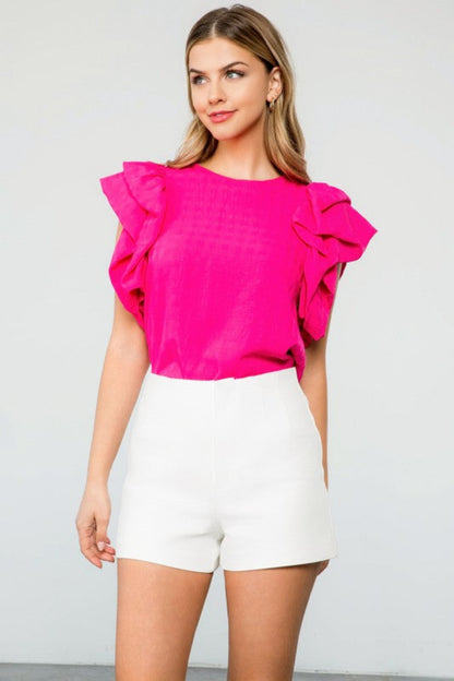 Woman wearing a bright pink blouse with ruffled sleeves and white shorts on a plain background