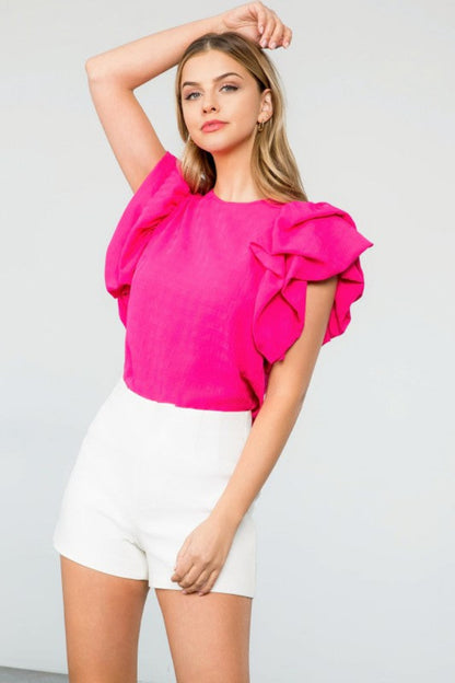 Woman wearing a bright pink top with ruffled sleeves and white shorts on a light gray background