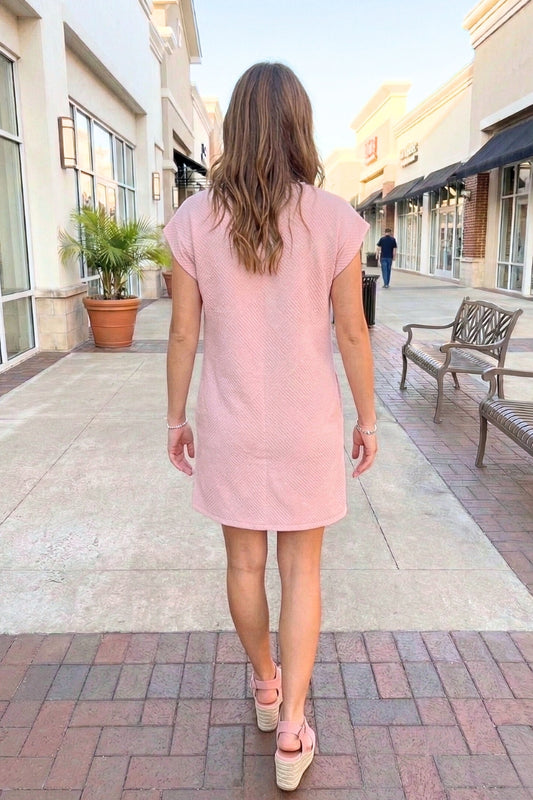 Woman in a pink dress walking on a sidewalk with buildings and benches in the background