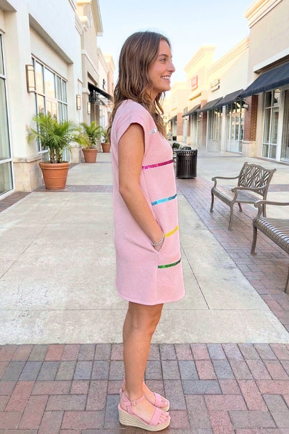 Woman in a pink dress standing on a sidewalk with buildings and plants in the background