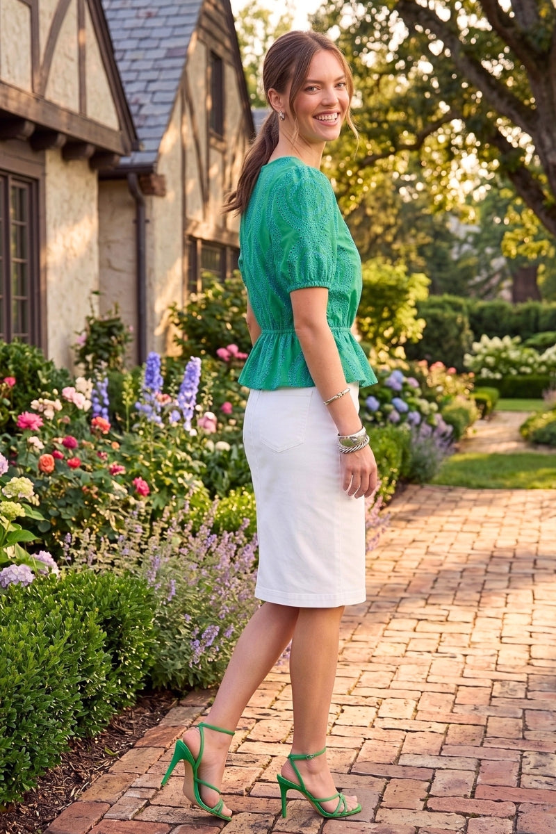 Woman in a green top and white skirt standing in a garden with flowers and a house in the background.
