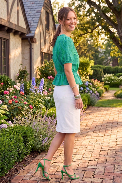 Woman in a green top and white skirt standing in a garden with flowers and a house in the background.