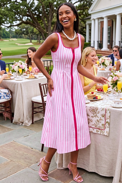 Woman in a pink and white striped dress standing outdoors at a social event with tables and people in the background.