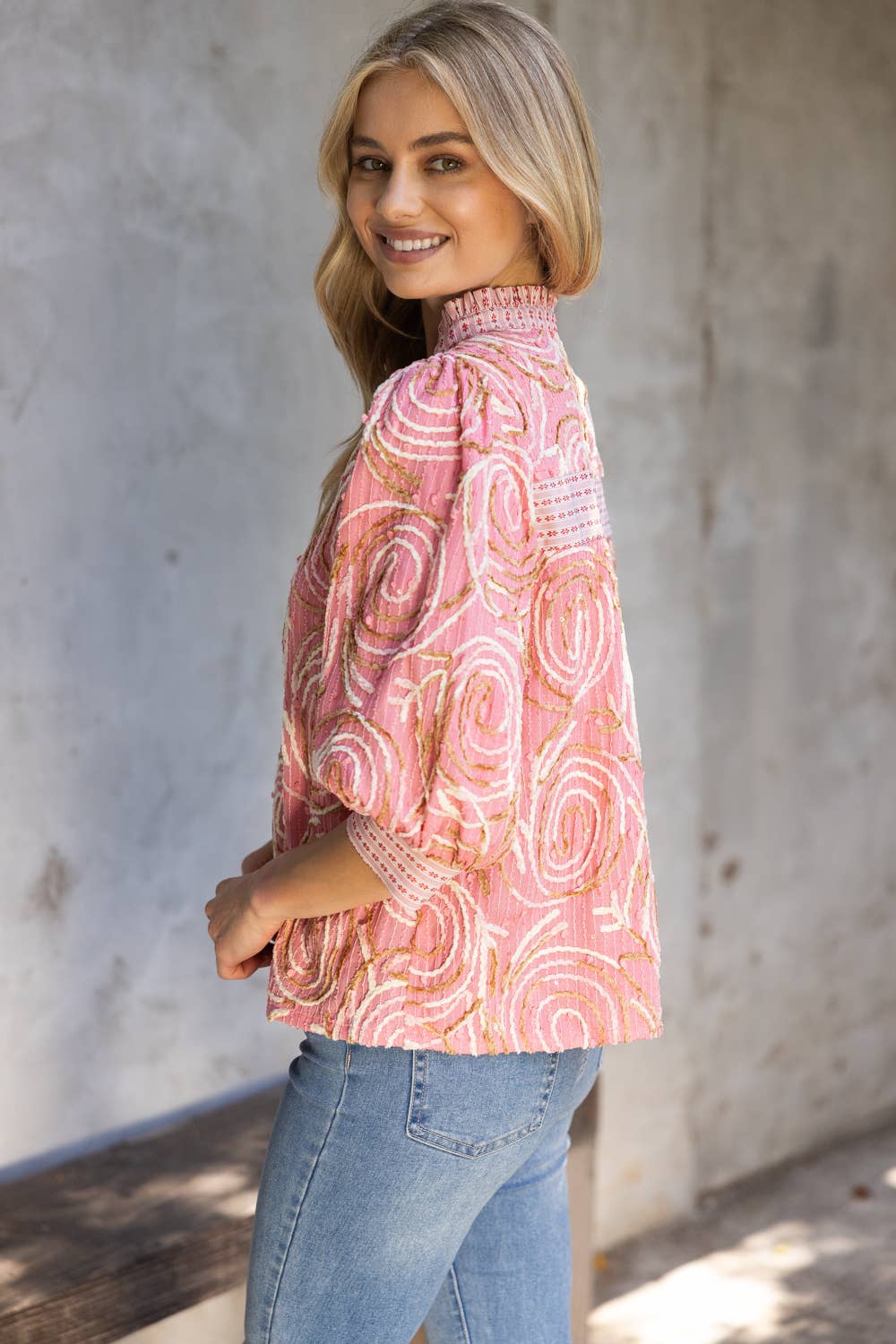 Woman wearing a pink patterned blouse and blue jeans against a concrete wall.