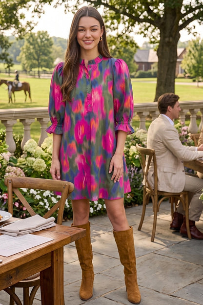 A woman stands outdoors on a stone patio wearing a short, loose-fitting dress with a bold pink, purple, and green abstract floral print, paired with tan knee-high boots. A man sits at a table in the background of a garden setting.