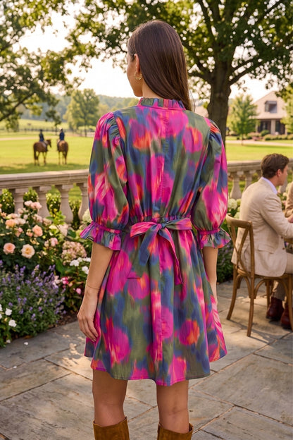 A woman viewed from behind wearing a short, tie-dye dress with pink, purple, green, and blue patterns. She is standing on a stone patio in an outdoor garden setting with a man seated at a table in the background.