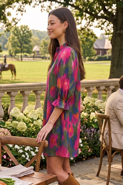 A woman wearing a colorful tie-dye short dress stands on an outdoor patio with garden flowers, a stone railing, and a horse visible in the background.