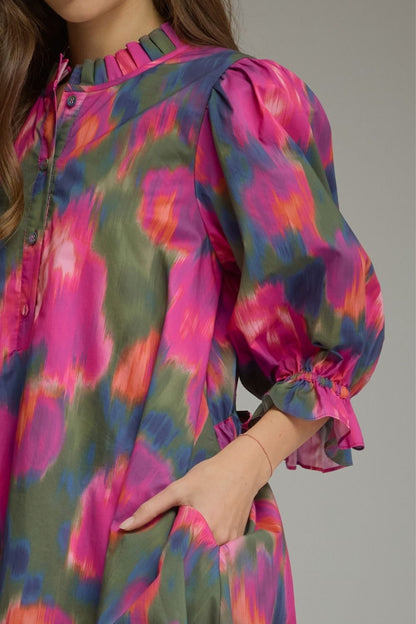 A close-up of a woman wearing a colorful abstract print dress with pink, green, and blue tones, featuring a ruffled high neckline, puff sleeves, and gathered cuffs.