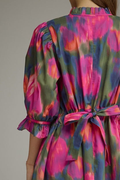 Close-up back view of a woman wearing a colorful tie-dye dress with puff sleeves and a self-tie waist belt. The dress features a vibrant pattern of pink, green, and orange on a neutral gray background.