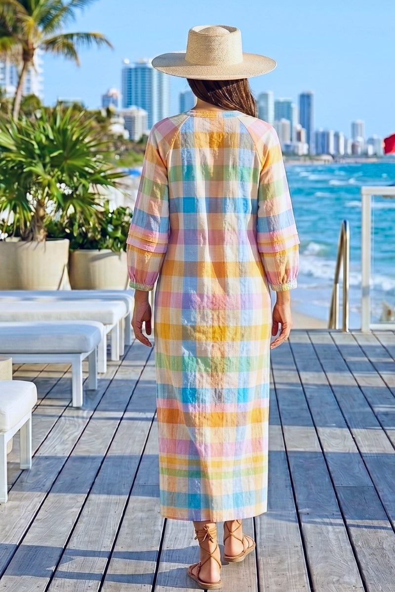 Person wearing a colorful checkered dress and straw hat by a waterfront with city skyline.