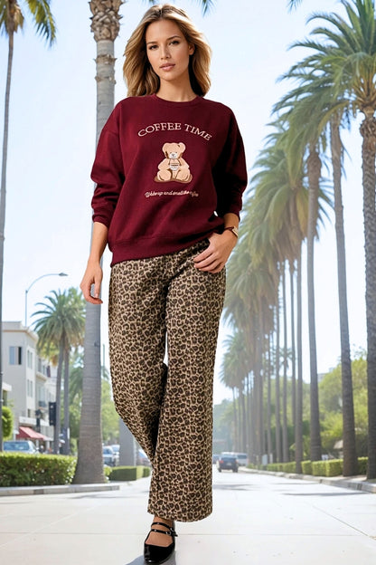 Woman wearing a maroon sweatshirt with a teddy bear graphic and leopard print pants on a palm tree-lined street.