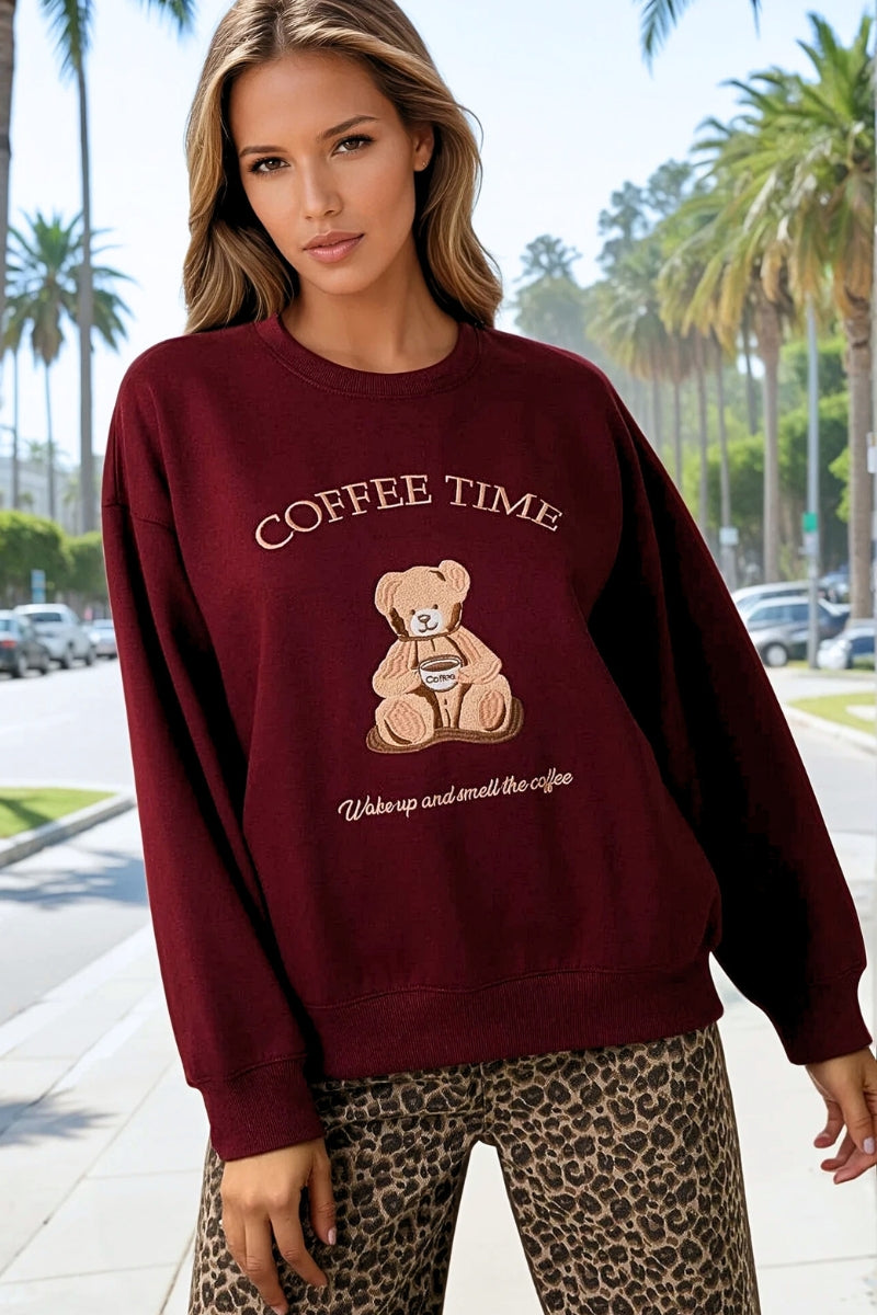 Woman wearing a maroon sweatshirt with a teddy bear graphic and 'Coffee Time' text, standing on a street with palm trees in the background.